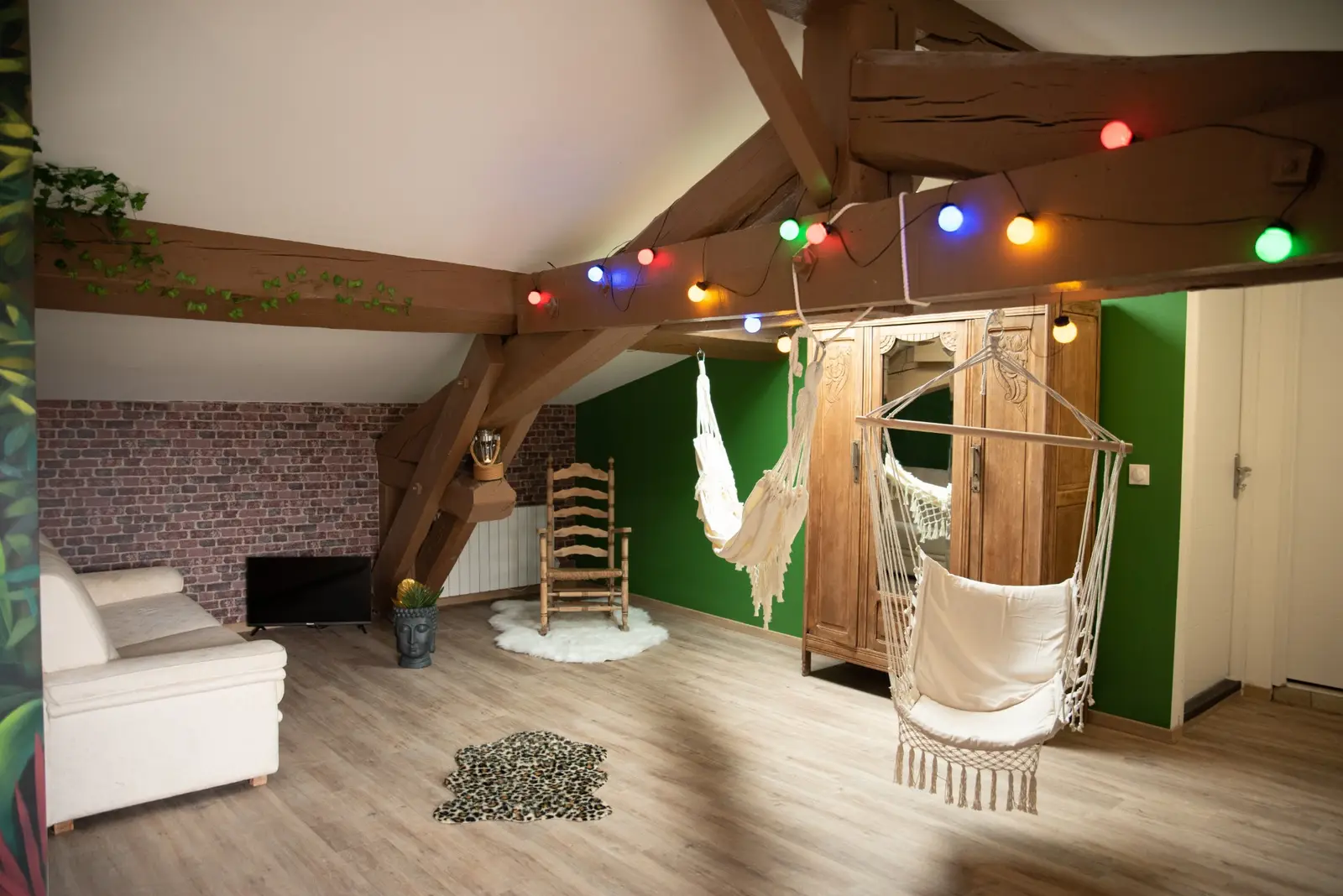 Salon sous les combles : poutres apparentes, fauteuils suspendus, guirlande colorée et mur vert
